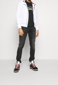 Chaqueta blanca con cremallera y detalles en rojo y azul marino, camiseta gráfica negra, vaqueros gris oscuro y botas negras y rojas con cordones.