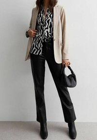 Donna che indossa un blazer beige, una camicetta a righe bianche e nere, pantaloni in pelle nera, stivali neri, mentre tiene in mano una piccola borsa nera.