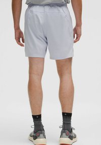 Homme portant un short de sport gris clair, des chaussettes noires à motifs et des chaussures de course grises, se tenant de dos face au spectateur.