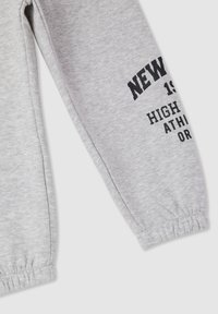 Pantaloni grigi in felpa con polsini elasticizzati, una stampa con la scritta "NEW YORK 19" e le parole "HIGH ATHLETIC OR" sulla gamba sinistra.