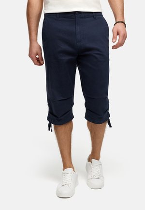 Mand iført marineblå knælange cargobukser med snore og hvide sneakers, stående mod en ensartet baggrund.