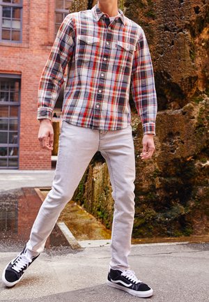 Mann trägt ein rotes, weißes und schwarzes kariertes Hemd, hellgraue Jeans und schwarze Turnschuhe, steht in der Nähe eines moosbewachsenen Felsens und eines Backsteingebäudes.