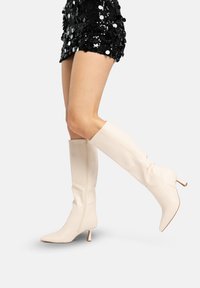 Benen met een glanzende zwarte minirok met pailletten en kniehoge roomwitte laarzen met hoge hakken en puntige neuzen op een lichte achtergrond.