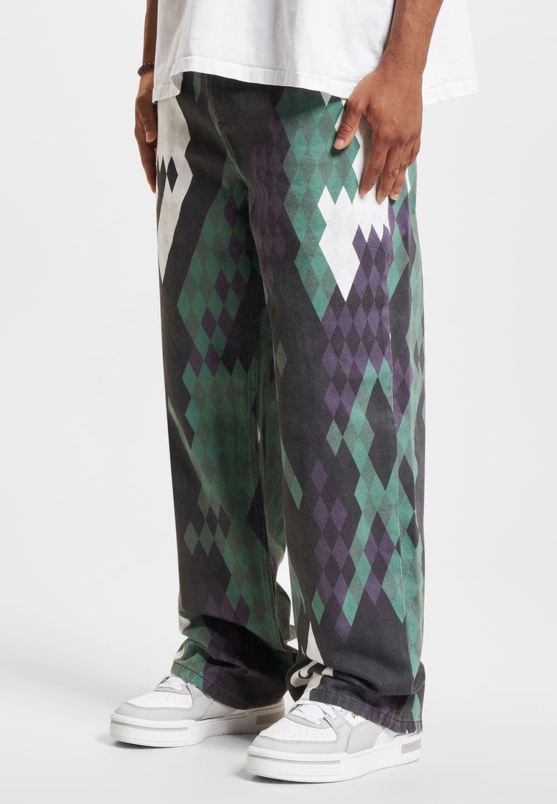 Pant legs feature a diamond pattern in green, purple, and black, with a loose fit. Paired with white sneakers and an oversized white shirt.
