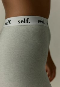 Heledad hallikas leggings, millega kaasneb valge elastne vöökoht, millel on must tekst "self." Pehme, veniv kangas, millel on ribaline tekstuur.