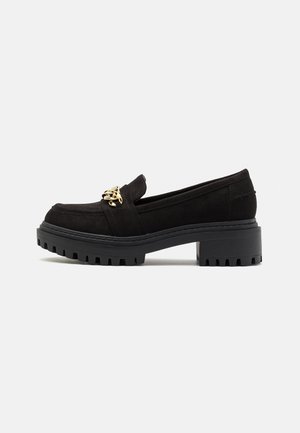 Mocassins en suede noir avec une semelle en caoutchouc épaisse et une texture striée, ornés d'une chaîne dorée à l'avant. Design à talon bas.