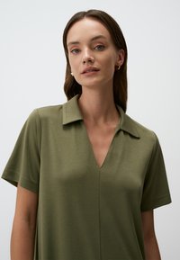 Polo shirt verde khaki con maniche corte, scollatura a V e tessuto liscio e strutturato. Design minimalista senza pattern o accenti visibili.