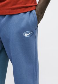 Άτομο που φορά μπλε φόρμα με λευκό λογότυπο Nike σε σχήμα γήινης σφαίρας στον μηρό, με το χέρι στην τσέπη και κόκκινο πουκάμισο ορατό.