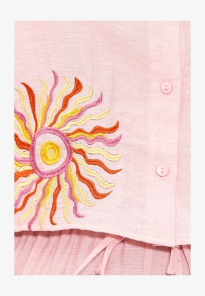 Tissu rose avec un design brodé de soleil présentant des vagues orange, jaunes et roses. Des boutons roses sur le vêtement ajoutent du détail.