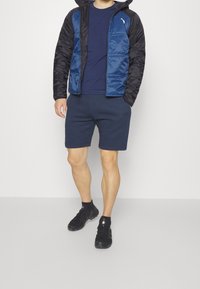 Marinblå och svart isolerad jacka med en slät yta, ihop med en marinblå t-shirt och matchande shorts, kompletterat med svarta skor.