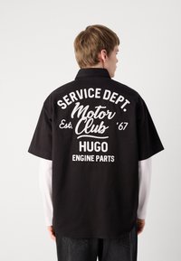 Zwart overhemd met korte mouwen met witte tekst: "SERVICE DEPT. Motor Club Est. '67 HUGO ENGINE PARTS." Klassieke kraag, losse pasvorm, van katoen.