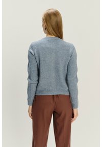 Jasnoniebieski sweter w miękkim, teksturowanym materiale, z długimi rękawami, ściągaczami przy mankietach i skróconym dołem, zestawiony z brązowymi spodniami.