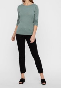 Maglietta a maniche lunghe, con texture, di colore verde chiaro, abbinata a jeans neri aderenti e tacchi aperti neri. I jeans hanno orli sfilacciati.
