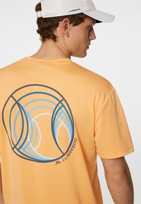 Orange kortärmad T-shirt med en blå och svart cirkulär grafik på baksidan. Tillverkad av mjukt, strukturerat tyg.