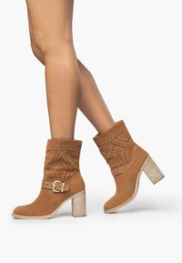 Beige Wildleder-Stiefeletten mit Blockabsatz. Mit dekorativen Ausschnitten und einem Schnallenakzent an der Seite. Glatte, weiche Textur.