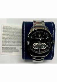 Zwarte roestvrijstalen horloge met een ronde wijzerplaat, zilveren accenten, drie sub-dials en een metalen band. Geplaatst op een blauwe fluwelen kussen.