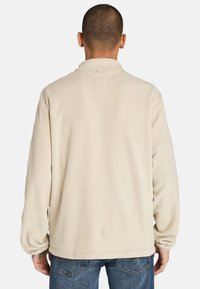 Beige fleecejacka med hög krage och långa ärmar. Har en etikett vid baksidan av halsringningen, slät textur och avslappnad passform.
