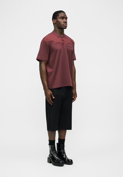 Άνδρας που στέκεται φορώντας μια μπουρνουζοκόκκινη κοντομάνικη πόλο μπλούζα, μαύρα σορτς μέχρι το γόνατο, μαύρες κάλτσες και λαμπερά μαύρα μποτάκια στον αστράγαλο σε λευκό φόντο.