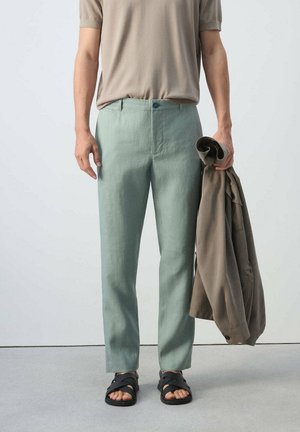 Mann trägt hellgrüne Hose, beige Kurzarmhemd, schwarze Sandalen, hält eine gefaltete braune Jacke in der rechten Hand.