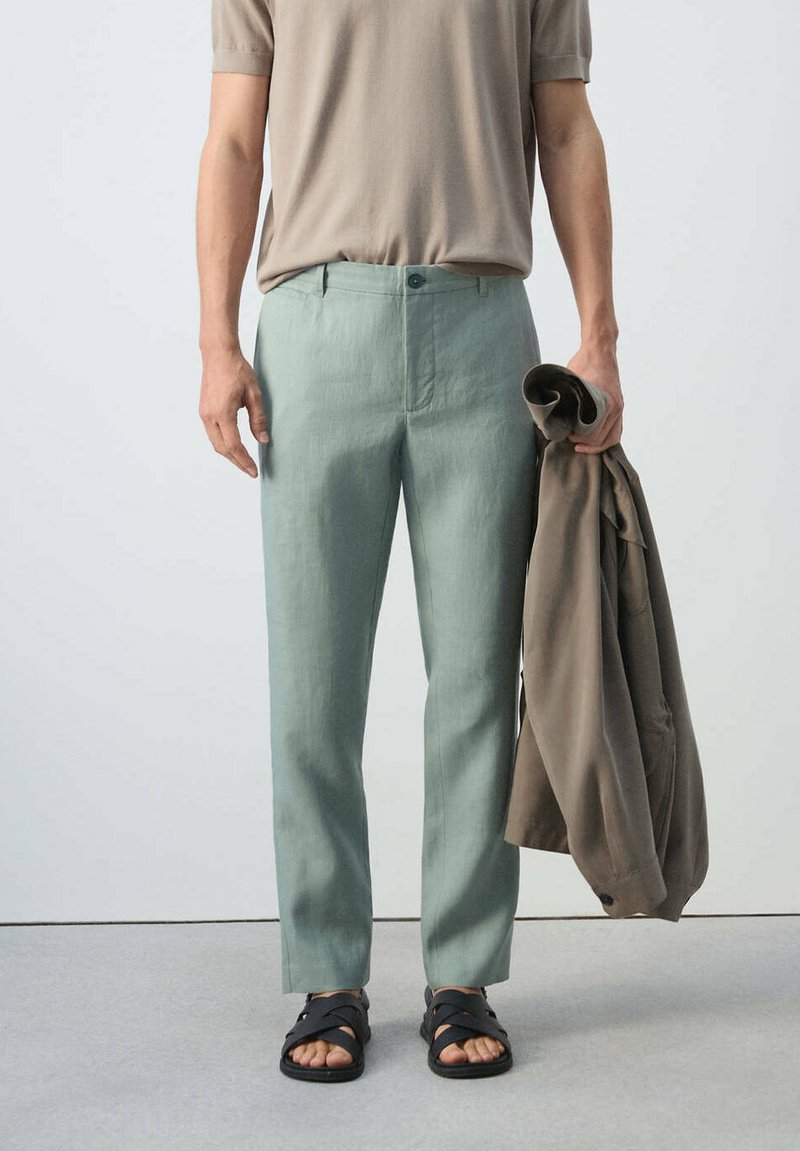 Homme portant un pantalon vert clair, une chemise beige à manches courtes, des sandales noires, tenant une veste brune pliée dans la main droite.