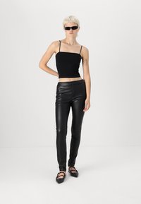 Top corto aderente nero con spalline sottili abbinato a pantaloni skinny neri simili al cuoio. Modella che indossa occhiali da sole neri piccoli e ballerine.