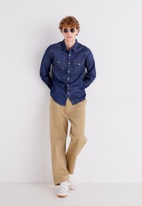 Camisa de mezclilla oscura con botones y dos bolsillos en el pecho, pantalones holgados beige y zapatillas blancas con detalles en naranja. Lentes de sol redondos. 