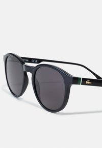 Ochelari de soare din plastic negru, cu o formă rotundă, lentile întunecate și un logo auriu pe braț. Accentuati cu verde pe templu.