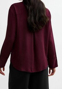 Burgundy long-sleeve blouse with a relaxed fit, smooth fabric, flat collar, and curved hem. Features a subtle back seam and rolled cuffs.