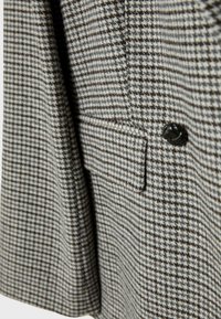 Manteau en motif pied-de-poule gris et crème avec un bouton noir, présentant une poche plaquée et une finition en tissu texturé.
