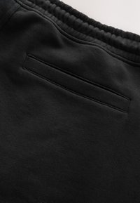 Pantalon de survêtement noir avec une ceinture élastique et une poche passepoil horizontale à l’arrière, montré en gros plan.