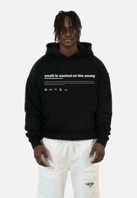 Fekete kapucnis sweatshirt, elülső zsebből, amelyen fehér szöveg olvasható: "a fiatalságot a fiatalokra pazarolják", valamint különböző logók alul.