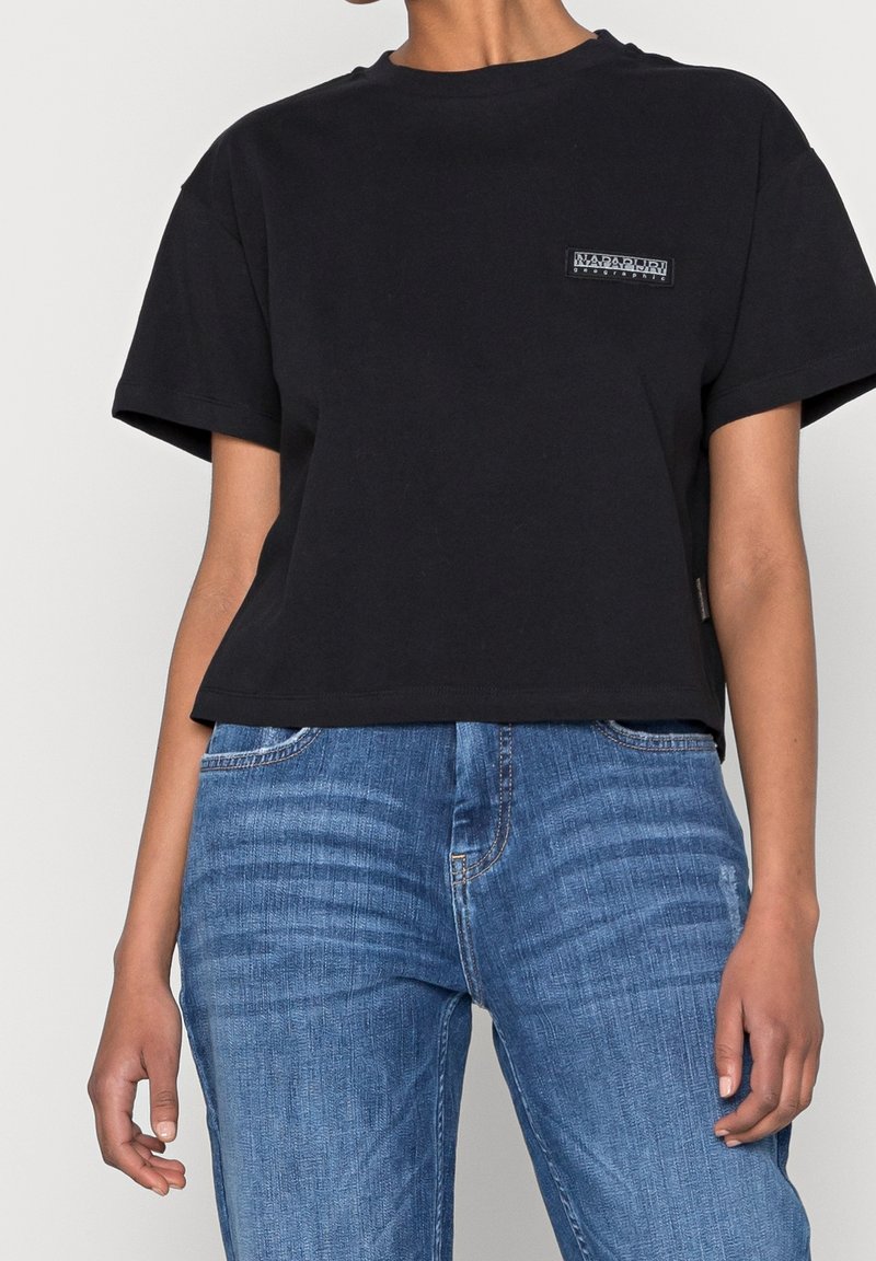 En person som bär en svart kortärmad croppad t-shirt med en liten bröstlogo och högmidjade blå denimjeans.