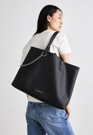 Kobieta w białej koszuli i dżinsach, niosąca dużą czarną torebkę typu tote marki Calvin Klein z detalem w postaci łańcuszka na ramieniu.