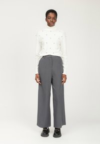 Mujer de pie con un suéter blanco de cuello alto con adornos, pantalones de pierna ancha grises y mocasines negros con cadenas doradas.