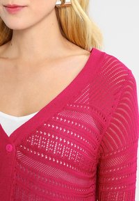 Fuchsia cardigan met een diepe V-hals, voorzien van ingewikkelde opengewerkte patronen en een enkele roze knoop. Gestructureerde stof met horizontale strepen.