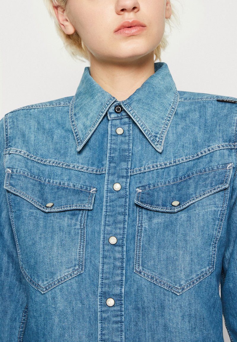 Denimhemd in hellblauer Farbe mit spitzem Kragen, Druckknöpfen und doppelten Brusttaschen mit kontrastierenden Nähte.