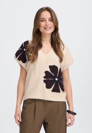 Femme portant un pull beige à manches courtes avec de grands motifs floraux foncés et un pantalon plissé marron, souriante, cheveux lâchés et un bracelet noir.
