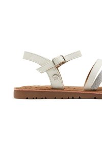 Sandalia de cuero blanca con dos correas cruzadas, hebilla ajustable y una plantilla texturizada; cuenta con una suela de goma marrón con patrón de zigzag.