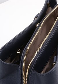 Sac fourre-tout en cuir bleu marine avec des garnitures en métal doré, comportant plusieurs compartiments zippés et un extérieur texturé. L'intérieur présente un imprimé de la marque.