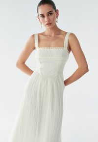 Balta, bezpiedurkņu kleita ar kvadrātveida izgriezumu, teksturētu audumu, mežģīņu apdari un plūstošu svārku. Elegants un viegls dizains.