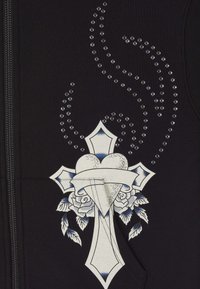 Giacchetto nero con zip caratterizzato da un design grafico di un cuore e una croce, abbellito da strass e dettagli floreali in bianco e blu.