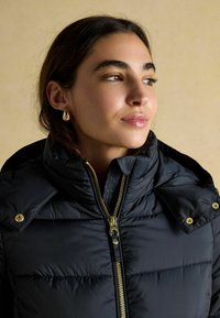 Mladá žena s tmavými vlasmi, ktorá má na sebe zlaté náušnice a čiernu prešívanú bundu s vysokým golierom a viditeľným zlatým zipsom a gombíkmi.