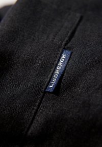 Zwarte stof met een gestructureerde afwerking, voorzien van een blauwe label met de tekst "LINDBERGH," gestikt nabij de naad.