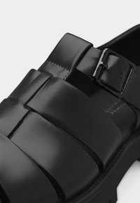 Μαύρο δερμάτινο σανδάλι με πλατιά λουριά και γυαλιστερή αγκράφα σε παχύ πάτο, εμφανιζόμενο σε κοντινή γωνία.