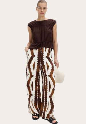 Donna che indossa una canotta marrone scuro arricciata, pantaloni bianchi a gamba larga con motivi geometrici marroni, che tiene un cappello bianco nella mano sinistra.