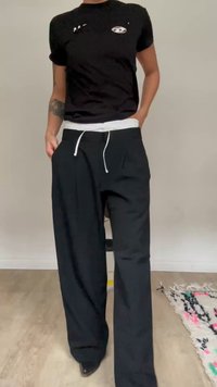 Osoba v černém tričku a volných černých kalhotách s bílým stahovacím šňůrkou stojí uvnitř na dřevěné podlaze.