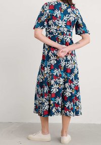 Robe midi florale bleue avec des motifs rouges, blancs, jaunes et verts, manches courtes, et jupe plissée, associée à des baskets de couleur claire.