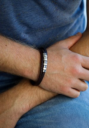 Brown braided leather bracelet with silver metallic accents, worn on a wrist, paired with blue fabric and denim background.