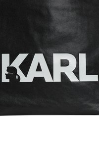 Borsa a tracolla nera con superficie texturizzata, caratterizzata da grandi testi bianchi che scrivono "KARL" e un design di silhouette sul lato sinistro.