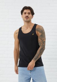 Canotta nera in cotone con scollo rotondo e un piccolo logo giallo sul lato sinistro del petto. Il modello ha dei tatuaggi visibili sulle braccia.